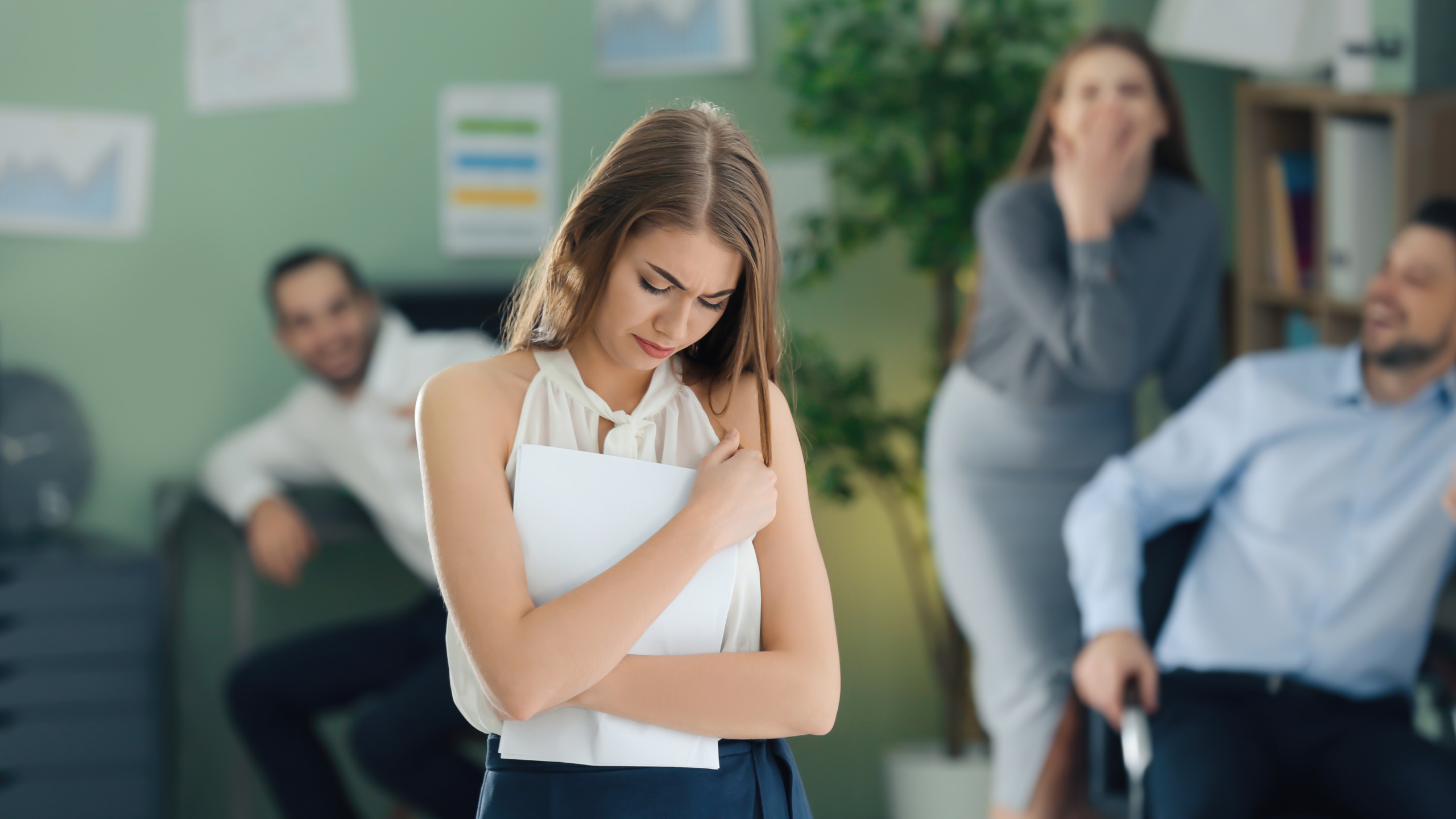 Upset female employee holding papers while coworkers in the background laugh in an office setting.